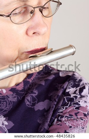 Middle aged woman with light skin and spectacles playing a silver flute against a gray background