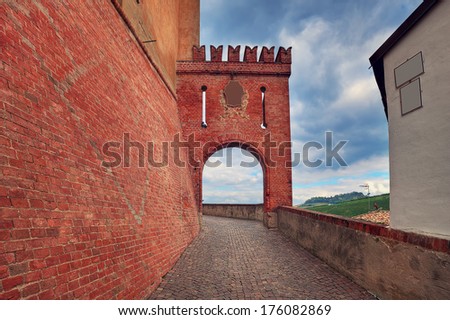 Narrow cobbled street along red brick wall and fragment of medieval arched passage in Barolo, Italy.