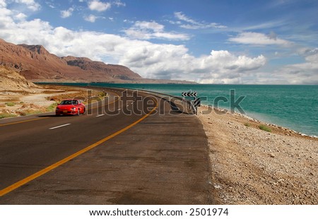 Red car on a highway that runs along the Dead Sea from one side and Edom Mountains at Arava Desert from the other in Israel.