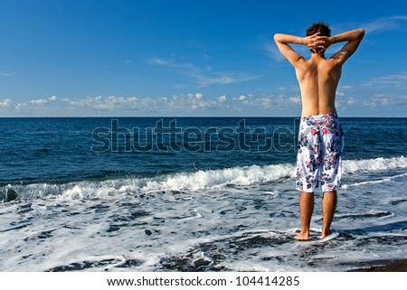 Ready for vocation. Young and healthy man wearing swimming trunks standing on the beach and looking at the sea.