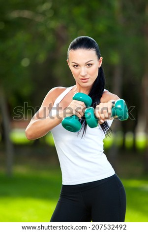Portrait of cheerful woman in fitness wear exercising with dumbbell, outdoors