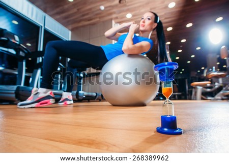 girl working out at the gym with a ball,  in the foreground of an hourglass , time concept to practice