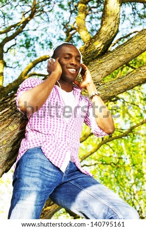 Young smiling african american standing on a tree branch in a park  and listens to music