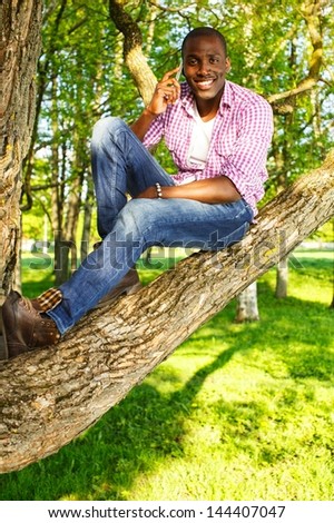 Young smiling african american sitting on a tree in park and calling with mobile phone