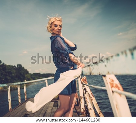 Stylish beautiful blond woman with white scarf standing near rails of old pier