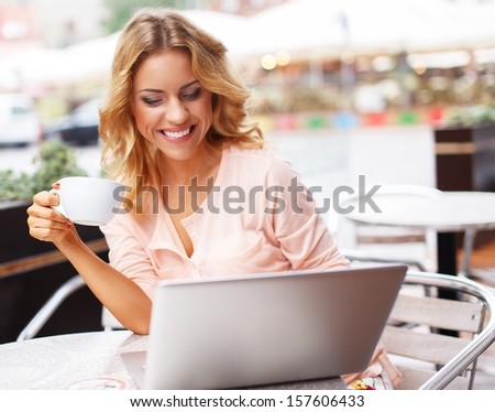 Smiling young woman with laptop and coffee cup alone in summer cafe