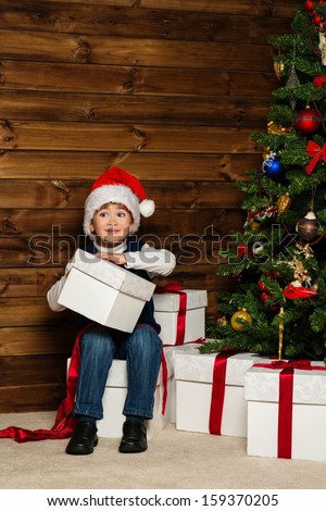 LIttle boy in Santa hat with gift box under christmas tree in wooden house interior