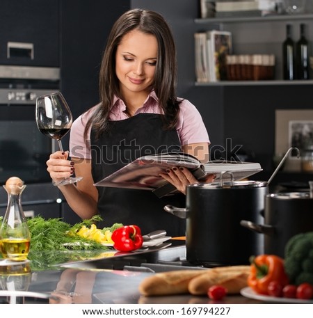 Cheerful young woman in apron on modern kitchen with cookbook and glass of wine