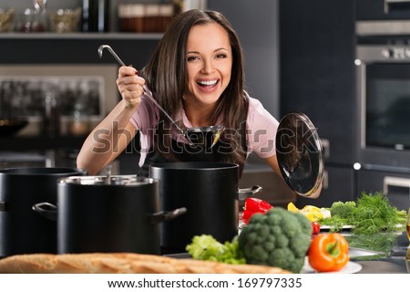Cheerful young woman in apron on modern kitchen will ladle tasting from pot
