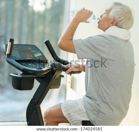 Senior Man Drinking While Doing Exercise On A Bike In A Fitness Club