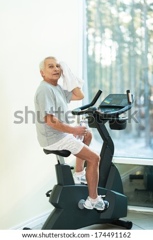 Tired senior man with towel on exercise bike in fitness club