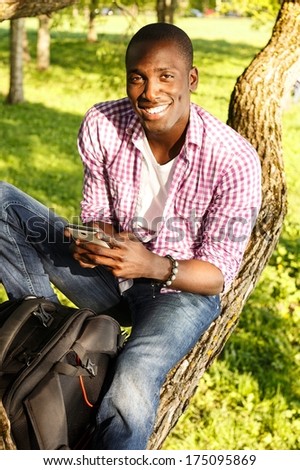 Young smiling african american sitting on a tree in park with mobile phone