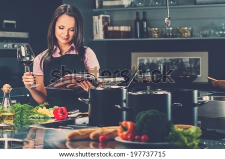 Cheerful young woman in apron on modern kitchen with cookbook and glass of wine