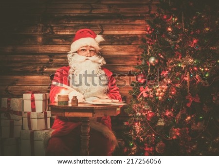 Santa Claus in wooden home interior sitting behind table and writing letters with quill pen