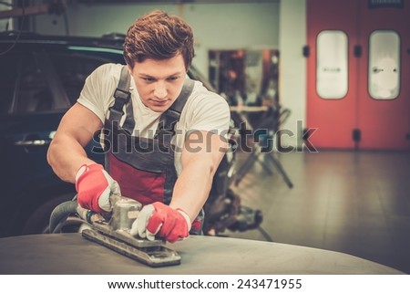 Young serviceman performing grinding with machine on a car bonnet in a workshop