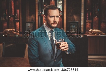 Confident well-dressed man with glass of whisky in luxury apartment interior.