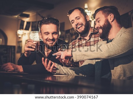 Cheerful old friends having fun with smartphone and drinking draft beer at bar counter in pub.