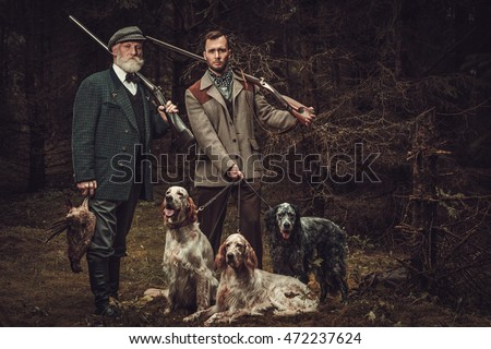 Two hunters with dogs and shotguns in a traditional shooting clothing on a dark forest background.
