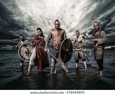 A group of armed Vikings, standing on the river shore with cloudy background.