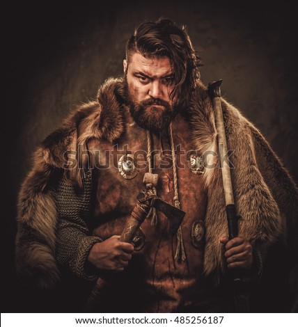 Angry viking with ax in a traditional warrior clothes, posing on a dark background.