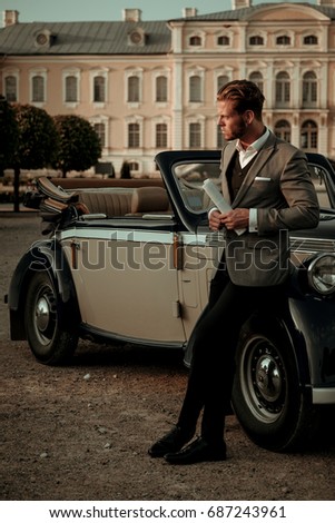 Confident wealthy young man with newspaper near classic convertible