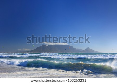 View of dramatic Table Mountain across Table Bay with big blue sky and shoreline waves in foreground.