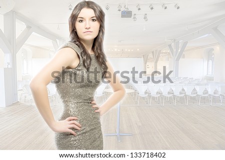 Beautiful young brunette woman with a curvy figure wearing an elegant silver dress standing in an auditorium with rows of seating, background faded and high key