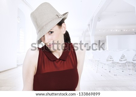 Beautiful glamorous woman in a red dress and trendy hat tipped at a rakish angle posing against a faded effect high key white architectural background in a spacious hall with rows of seating