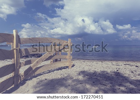 Old rustic wooden fence on a beach leading down towards the calm ocean with a yacht visible in the far distance under a cloudy blue sky