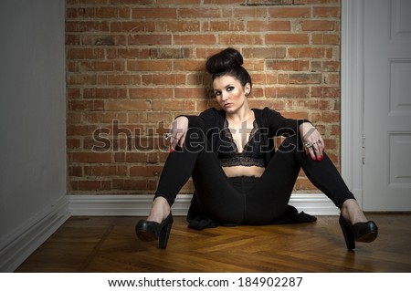 Moody beautiful elegant woman in a fashionable stylish black outfit sitting on a parquet floor with her legs spread looking at the camera with a sultry speculative expression