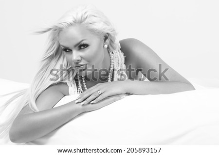 Beautiful glamorous blond woman wearing pearl necklaces and jewellery relaxing on her stomach on a bed looking sideways at the camera with a serious expression, greyscale portrait