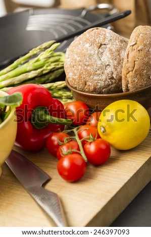 Healthy assortment of fresh vegetables including red bell pepper, lemon, asparagus tips and tomatoes with wholegrain rolls on a wooden kitchen counter with a sharp knife