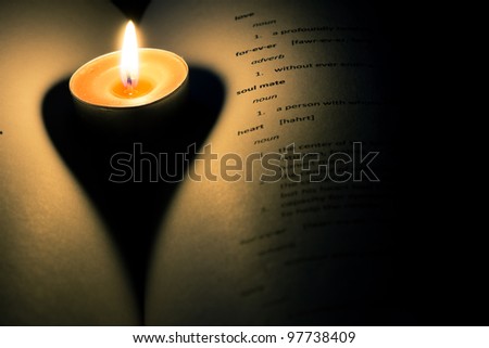A candle in the crease of a book casting a heart shaped shadow.  There are definitions of various love related words on the page.