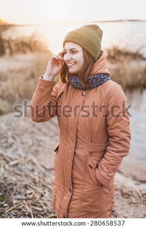 Young smiling girl in a green hat and jacket stand near the river in the sunset light. Woman\'s portrait outdoors. Lifestyle. Soft focus