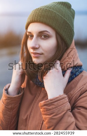 Young attractive girl in a green hat and jacket near the river in the sunset light. Close-up woman\'s portrait outdoors. Lifestyle. Soft focus