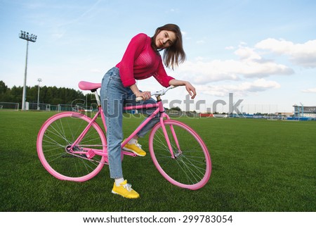 Young sexy woman standing beside bicycle on the football field. Sexy girl in a pink blouse hipster and cycling. Girl and bike fixed