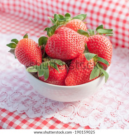 Small bowl filled with succulent juicy fresh ripe red strawberries on table with red checkered tablecloth and lace