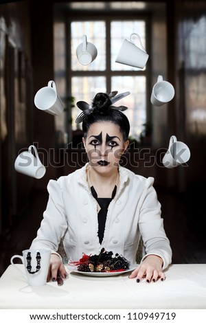 Young female with pierrot style makeup is sitting near the table with cups flying around her