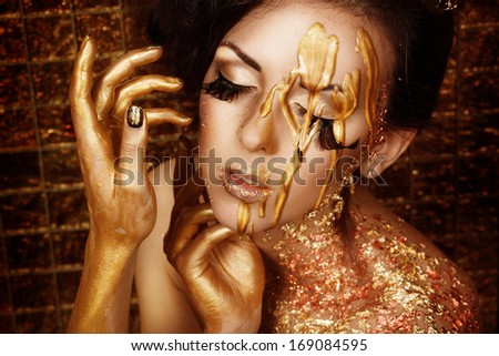 Closeup portrait of beautiful young brunette with long feather lashes in gold liquid paint and bronze foil on golden background holding her hands near her face