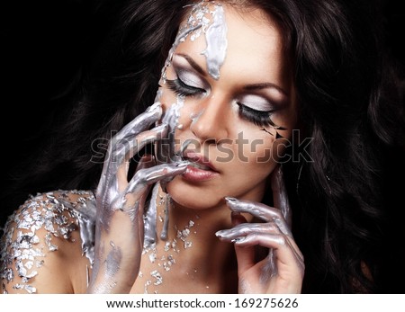 Closeup portrait of beautiful young brunette with long feather lashes in silver liquid paint and  foil on black background holding her hands near her face