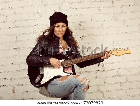 Young teenage looking woman playing her black electronic guitar wearing her leather jacket and knitted beanie standing in front of white brick wall