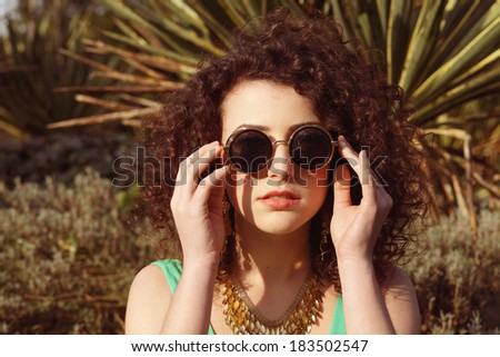 Young female in tropical garden is wearing round sunglasses and holding them near her face