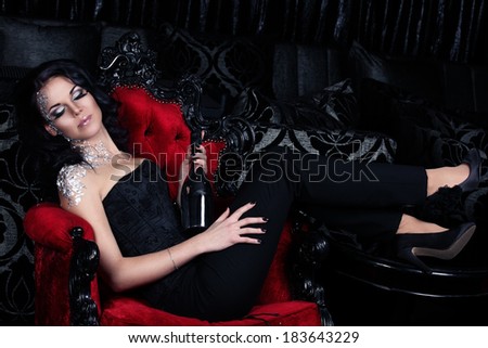 Attractive woman with silver makeup is having a party and holding silver champange bottle in her hands sitting on the red vintage gothic chair