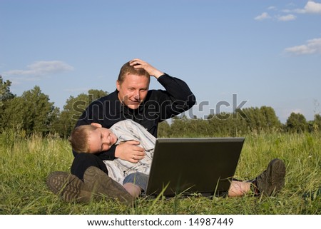 The daddy and the son play the laptop on a green grass