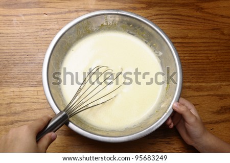 Child hands mixing some egg, milk, sugar, with a whisk in a metallic bowl on wooden table