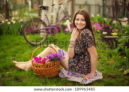 Beautiful young woman in dress sitting on grass with basket of flowers and old vintage bicycle