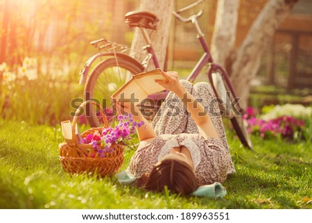 Beautiful young woman in dress laying on grass with basket of flowers and reading book near old vintage bicycle