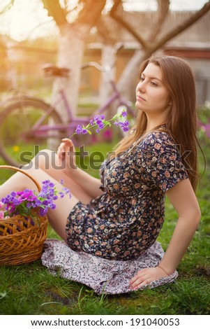 Beautiful young woman in dress sitting on grass with basket of flowers and old vintage bicycle