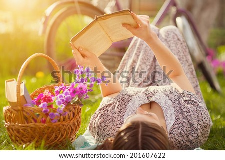Beautiful young woman in dress laying on grass with basket of flowers and reading book near old vintage bicycle
