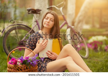 Beautiful young woman in dress sitting on grass with basket of flowers and reading book near old vintage bicycle